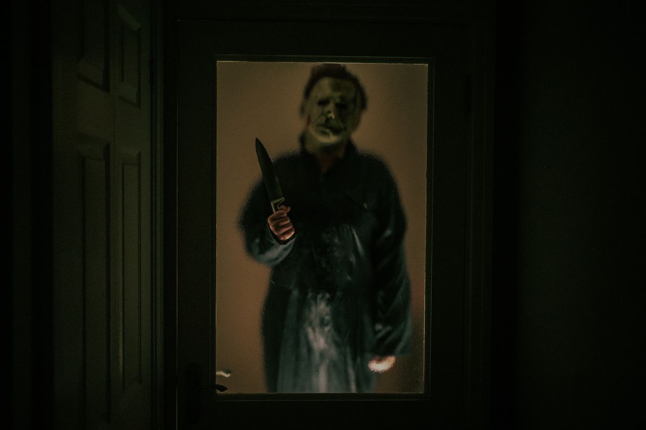 A spooky masked figure stands outside a dimly lit door, creating a chilling Halloween ambiance.