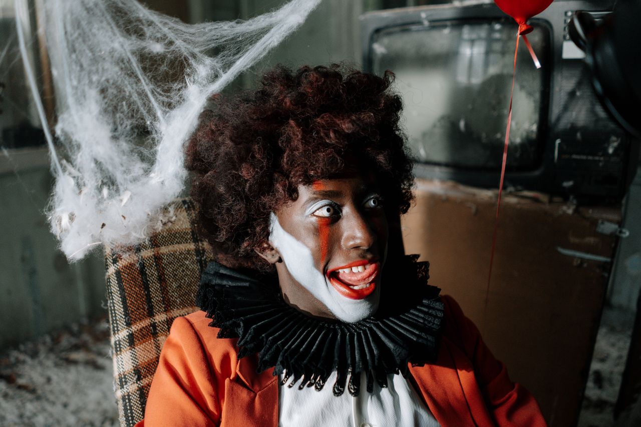 Dreadful clown with big smile and red balloon in eerie, cobweb-filled room.