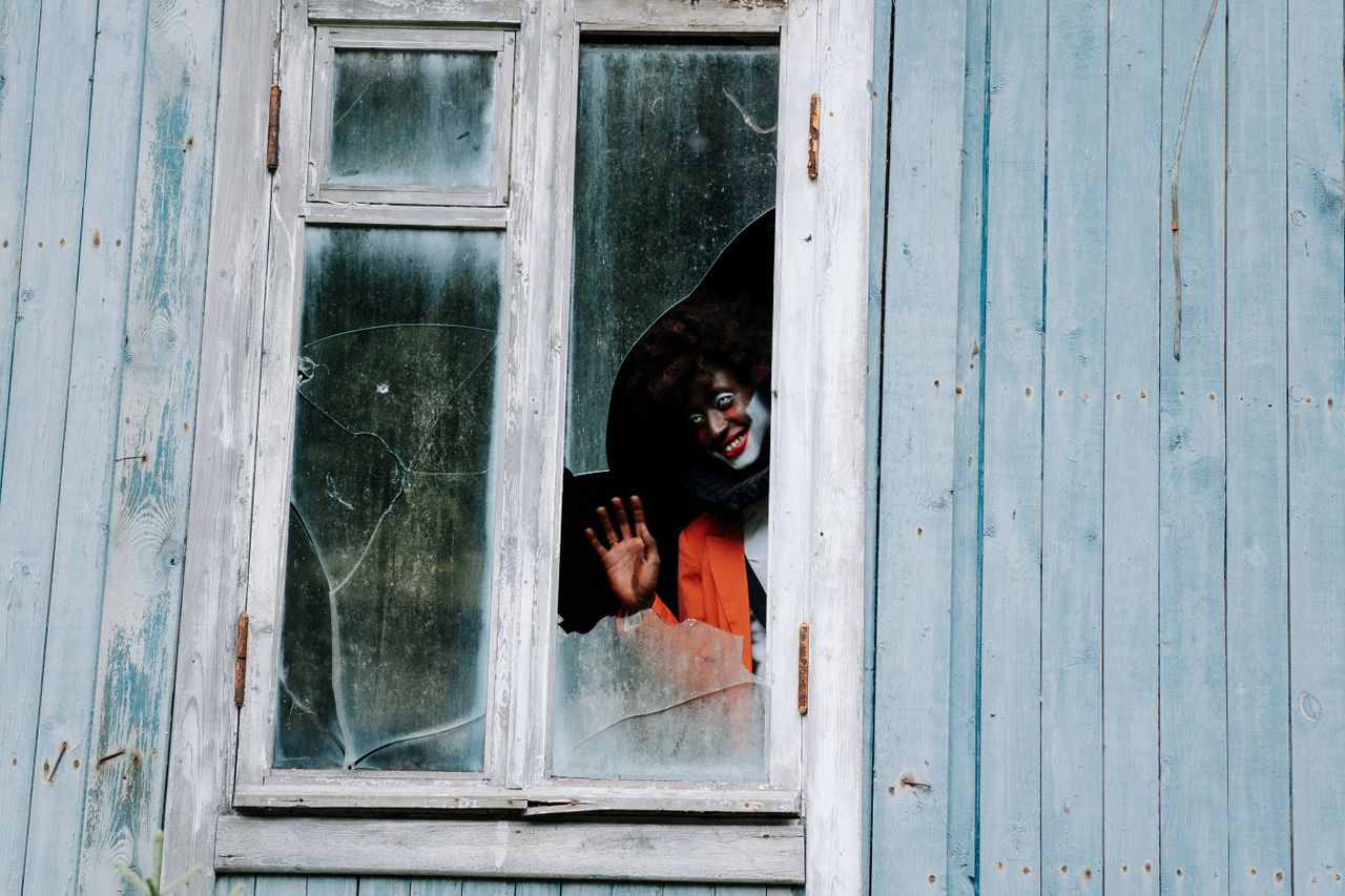 A terrifying clown peeks through a broken window of an abandoned house, exuding eerie horror vibes.